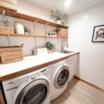 A cozy small laundry room features a white washer and dryer, wooden shelves with baskets, and a countertop with a sink. Decor is complemented by framed photos and plants, making efficient use of every inch.