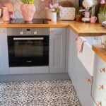Small gray shaker galley kitchen with butcher block counters, copper cabinet knobs and faucet, a white herringbone subway tile backsplash, patterned black and white encaustic floor tile, a farmhouse sink, pink tulips in vases on the counter, and a stainless Bosch oven.