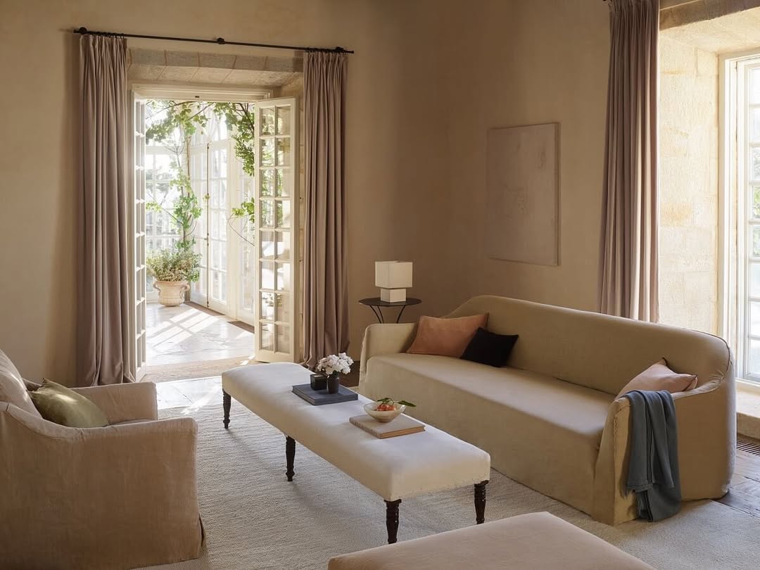 Beige slipcovered sofa with a coral and plum pillow and a long upholstered bench coffee table in a small warm neutral living room with open French doors to a planted courtyard