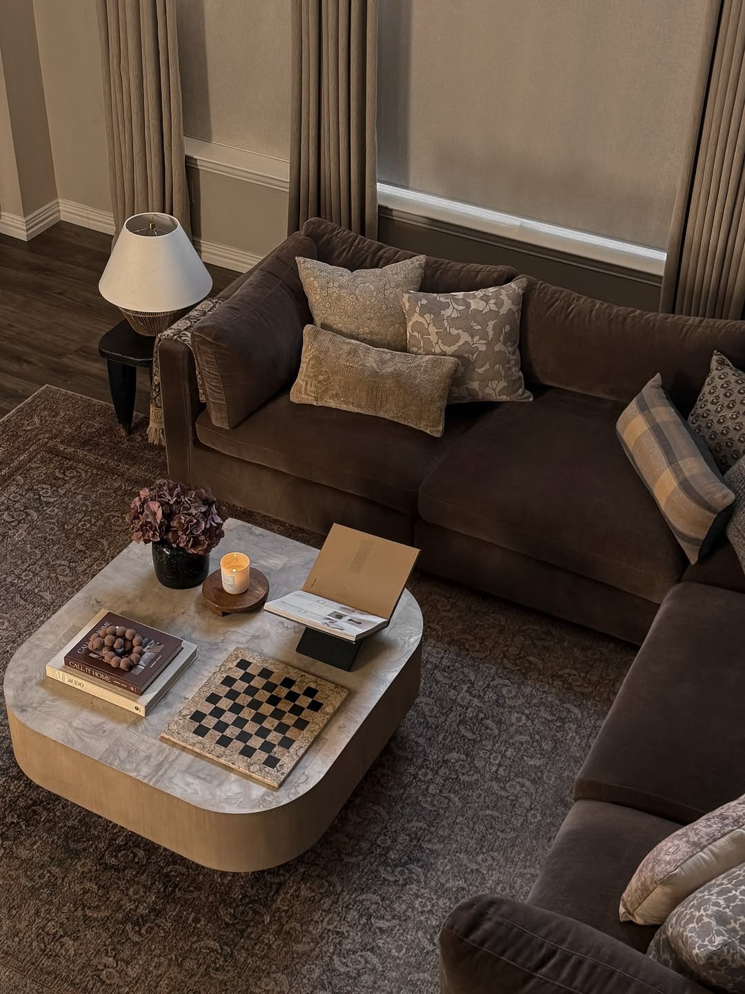 Dark chocolate brown velvet sectional with layered patterned pillows and a round marble-top coffee table on a wood base holding a candle a hydrangea and a checkerboard in a moody small living room