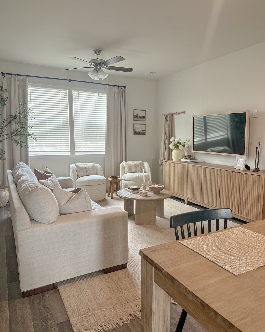 Cream fabric sofa with two boucle swivel chairs facing a long fluted light wood TV console with white tulips in a bright small living room with layered neutral rugs and a round wood coffee table