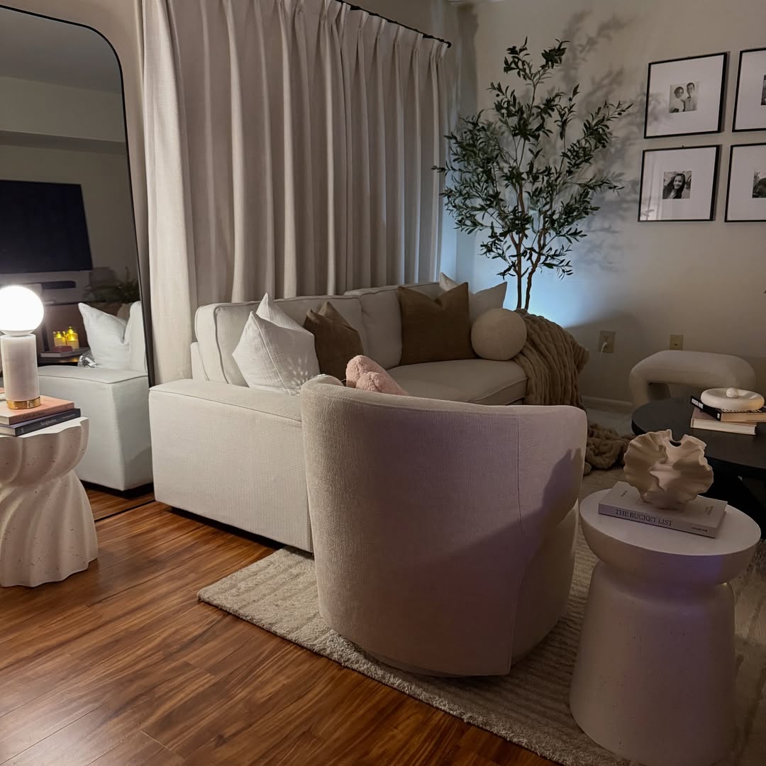 Cream boucle swivel chair facing a cream sofa with brown and white pillows beside a tall olive tree lit by a mushroom-shaped globe lamp in a cozy small living room at night with a tall arched mirror