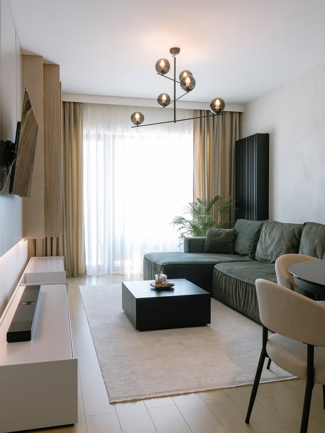 Deep green velvet sectional with a low square black coffee table under a six-globe black metal chandelier beside a tall palm plant in a small modern living room with taupe curtains