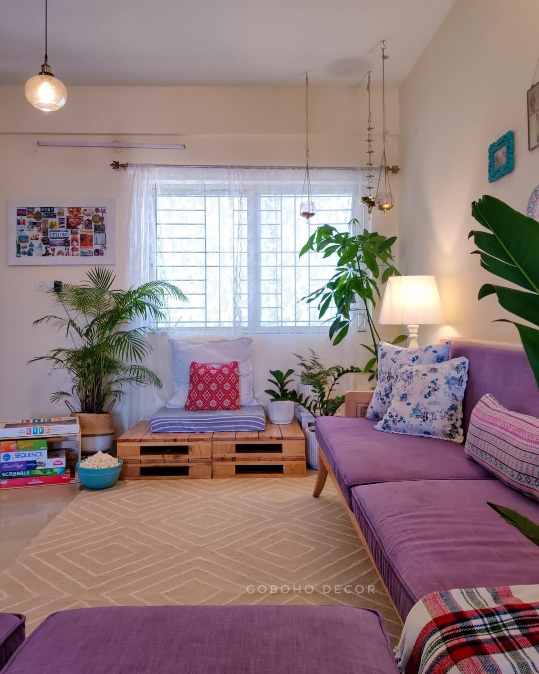 Purple velvet sofa with a blue floral pillow and tribal pattern pillow framed by a tall palm and bird of paradise plants with a pallet-wood side bench and a glass pendant light in a boho small living room
