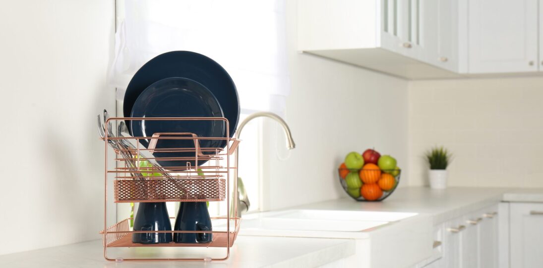 Rose gold copper wire dish rack sitting on a bright white kitchen counter beside a farmhouse sink, holding navy blue plates leaning at the back, upside-down navy mugs on the bottom tier, and stainless cutlery in the side caddy, with a black wire fruit bowl and small potted plant softly blurred in the sunlit background.