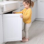Small girl with pigtails in a yellow floral cardigan and pale pink sneakers pulling open the door of a white under-sink cabinet in a bright kitchen with a farmhouse sink, brushed nickel handles, and light wood floors.