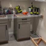 Walk-in pantry mid-renovation with white wire shelves along the back and side walls, two gray shaker base cabinets pushed against the back wall, cordless power tools sitting on the wire shelf, patched drywall spots, and exposed wood framing on the tile floor.