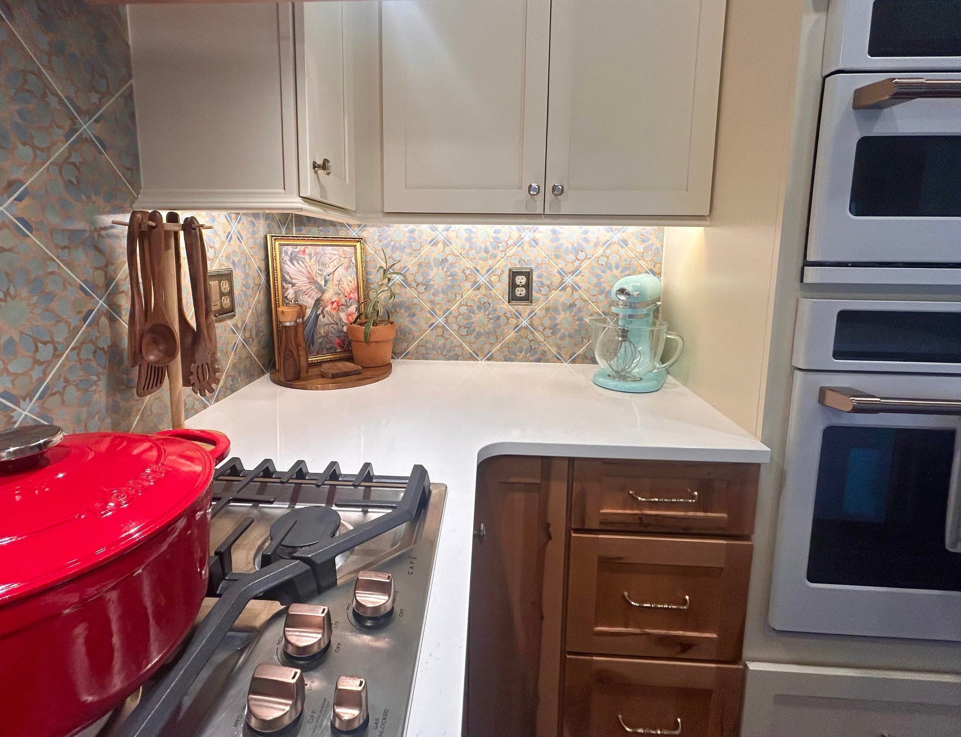 Corner view of the Victorian kitchen cooktop with the Moroccan tile backsplash, a mint green KitchenAid mixer on the counter, hanging wooden spoons, and the white double wall ovens built into the alder cabinets.