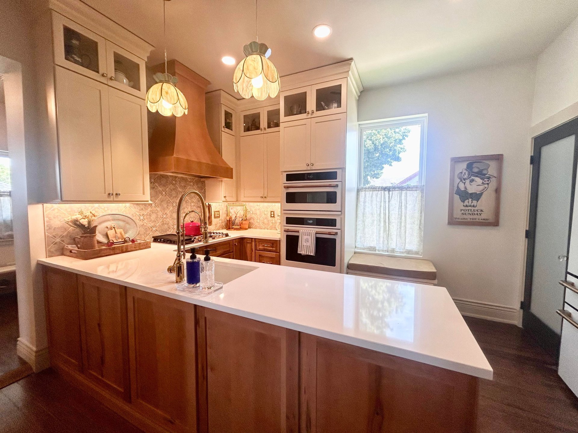 Wide view of the remodeled Victorian kitchen showing the copper range hood, stained glass tulip pendants, cream and wood cabinets, Moroccan backsplash, and a cushioned window seat under a bright window where the old refrigerator used to stand.