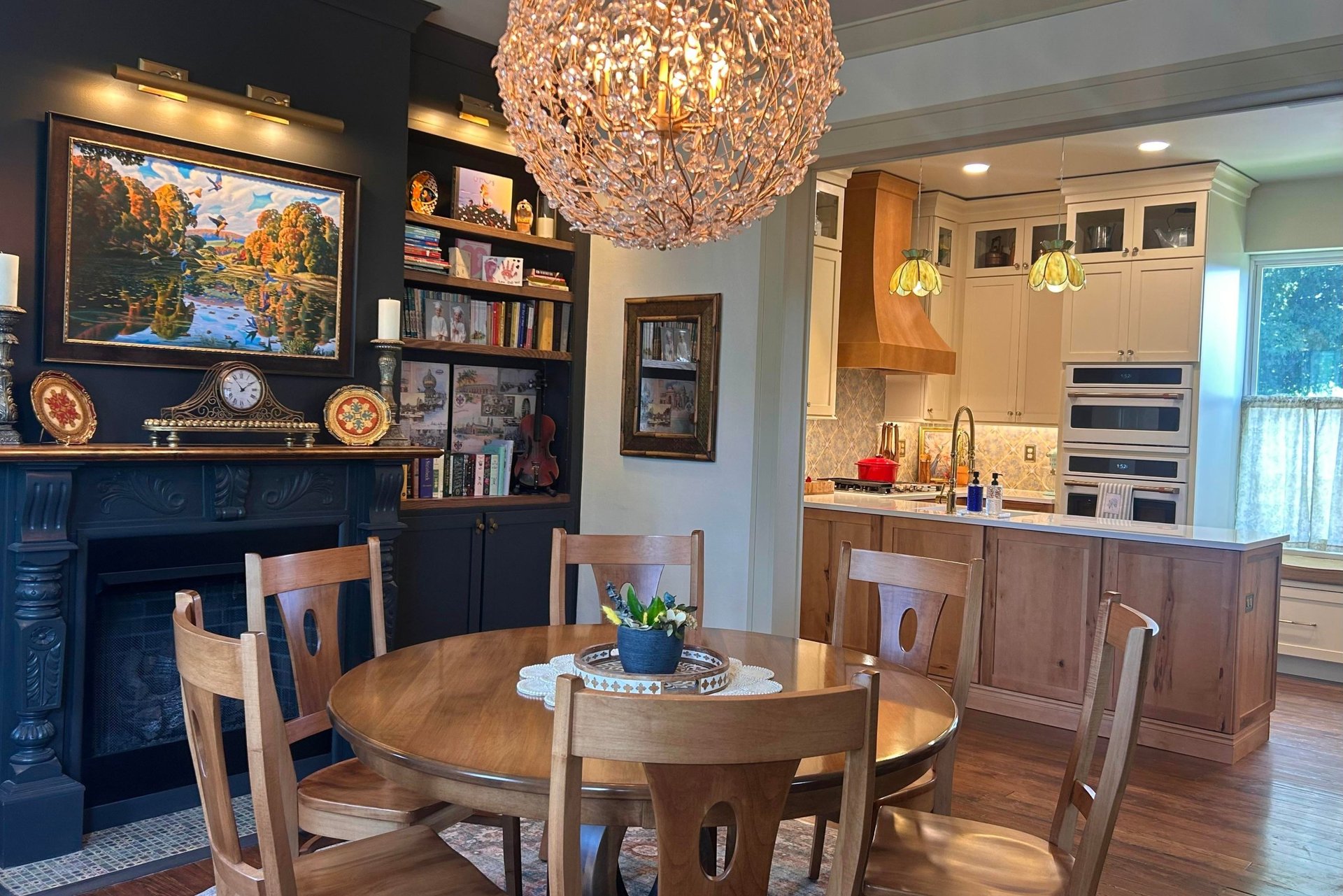 Dining nook off the Victorian kitchen with a round wood table, carved black fireplace with mantel art, dark painted bookshelves, and a large crystal branch chandelier overhead, with the new kitchen visible through the archway.