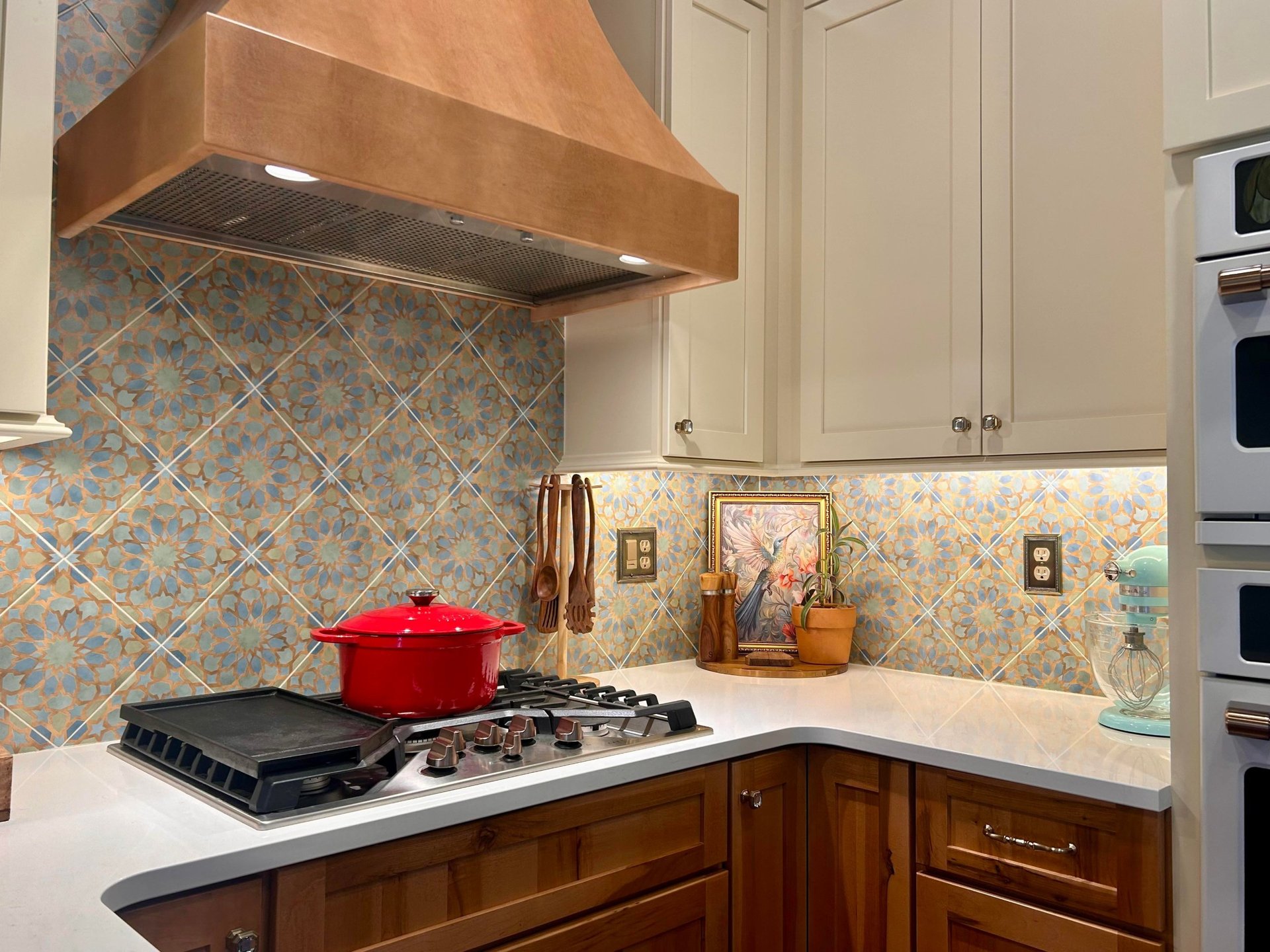 Hand-painted Moroccan-pattern tile backsplash in blue, terracotta, and cream behind a stainless gas cooktop with a bright red Dutch oven and a copper range hood above.