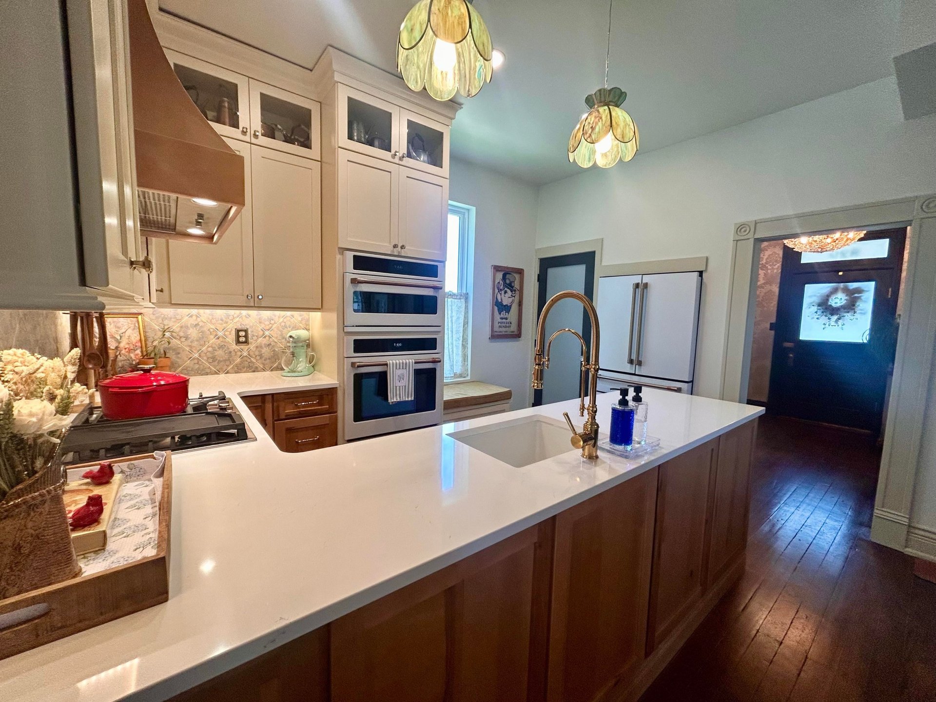 Long view across the quartz peninsula with brass gooseneck faucet toward the white French-door refrigerator in its built-in closet and the front door of the Victorian home beyond.