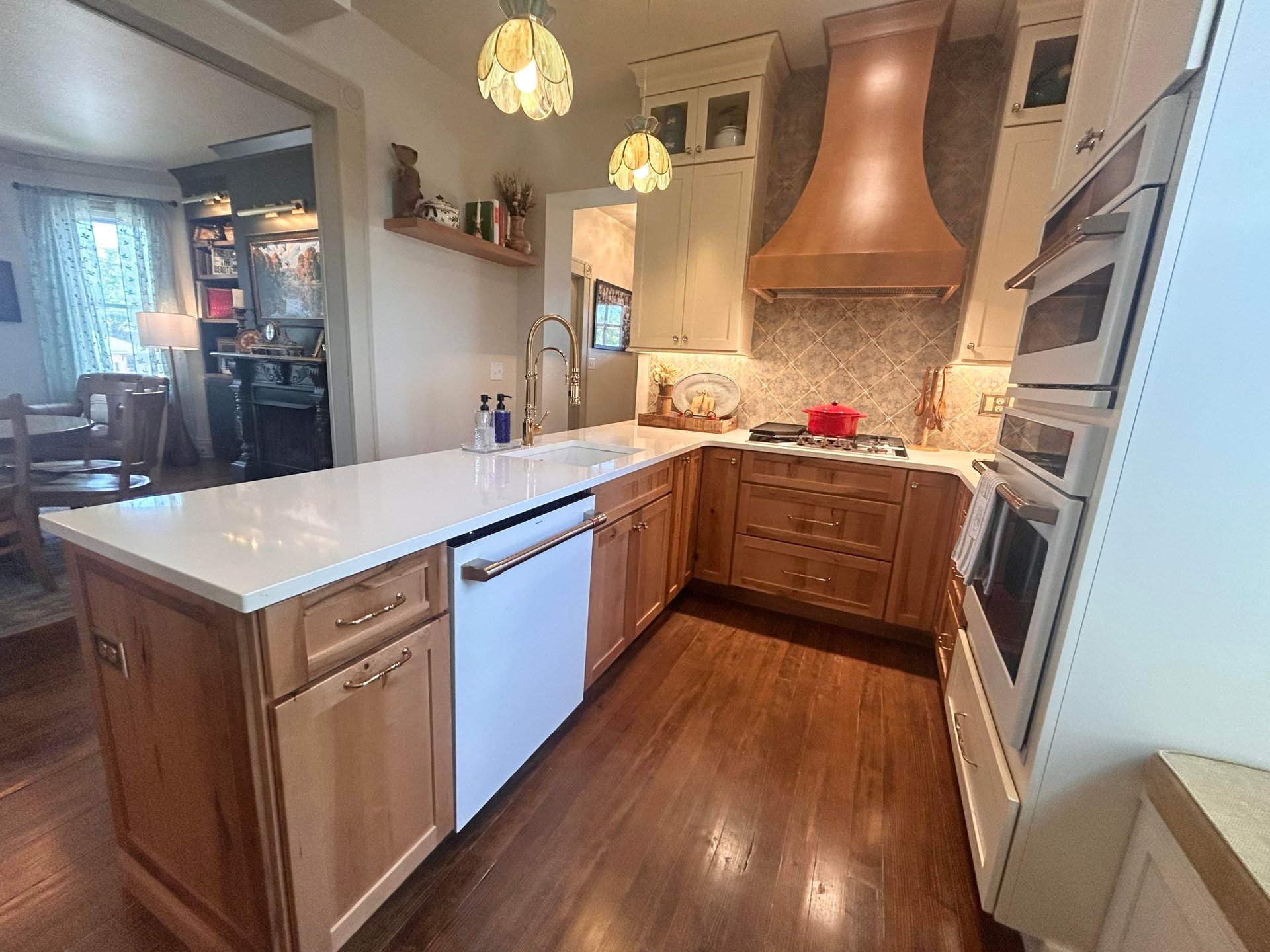 Angled view of the remodeled Victorian kitchen with cream upper cabinets stacked to the ceiling, warm stained wood lower cabinets, a copper range hood, white GE Cafe double wall ovens with brushed bronze trim, and the Moroccan backsplash.