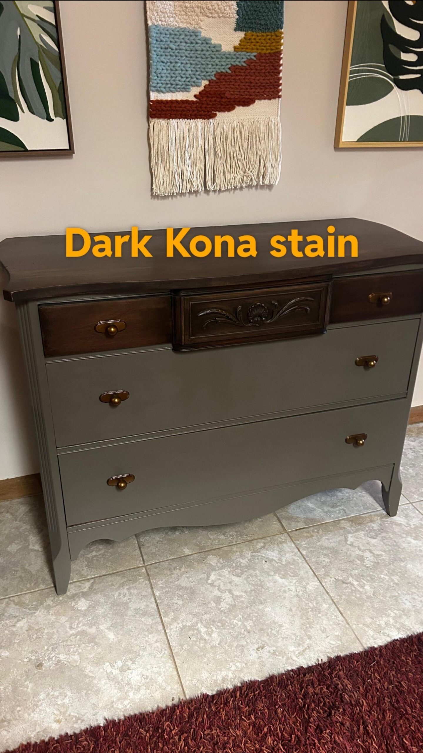 Refinished 1940s bow-front dresser with an olive-gray painted body, carved center drawer and top finished in a near-black Kona stain, warm brass pulls, styled under boho wall art and a woven wall hanging.