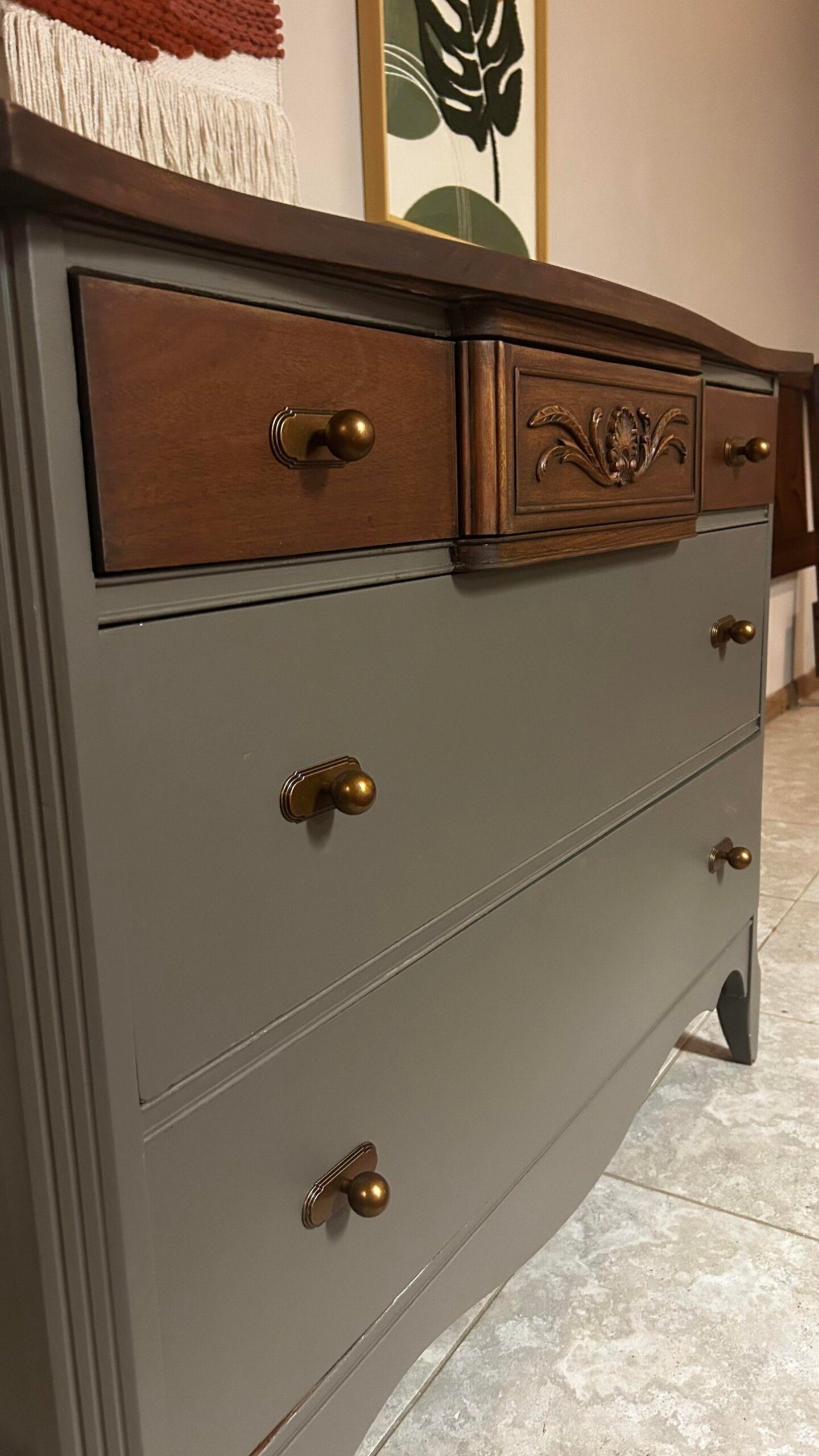 Angled side view of the dresser showing the warm walnut top catching the light next to the olive-gray body, carved center drawer, and brass hardware.