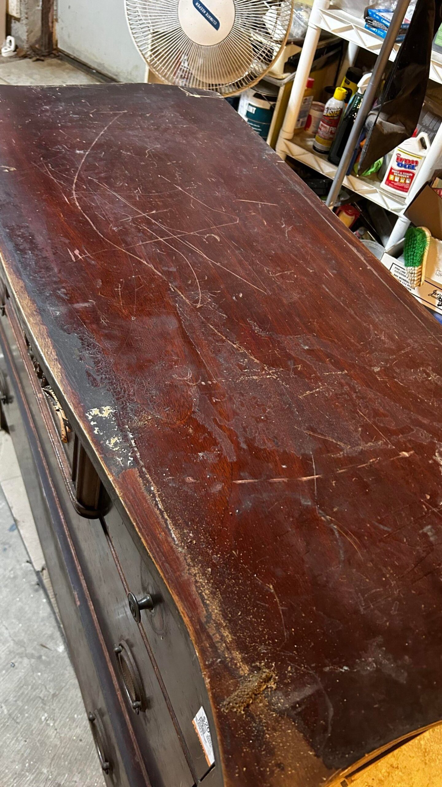 Close-up of a dark mahogany 1940s dresser top covered in deep scratches, white water rings, black burn marks, and raw wood where the finish has worn off, photographed in a basement workshop.
