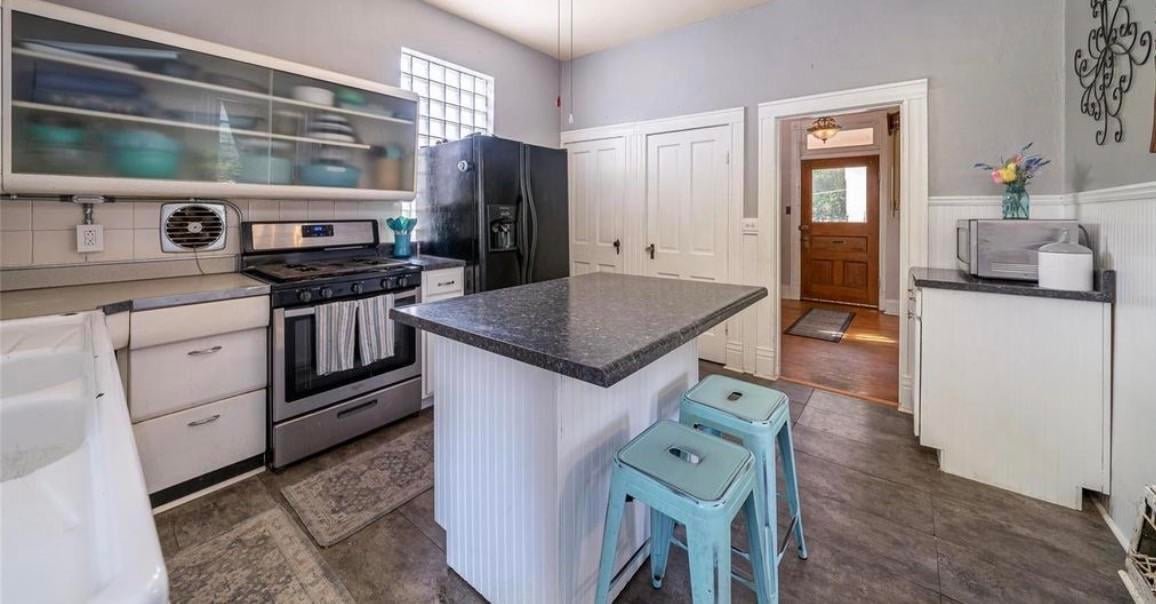 Before photo of a 1960s remodel in a Victorian kitchen showing a black side-by-side refrigerator blocking a glass block window, white laminate counters, and teal painted metal bar stools.