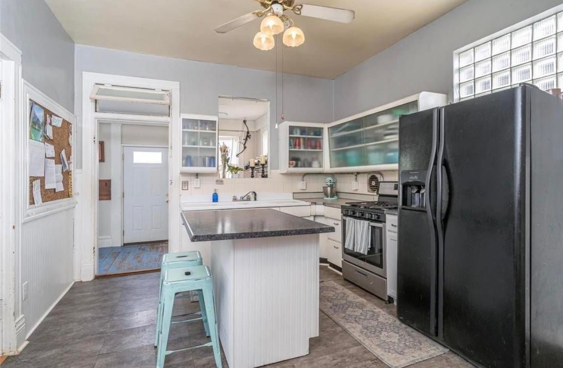Second before view of the old kitchen showing frosted glass sliding upper cabinets, a brass ceiling fan with globe lights, a small window above the back door, and a lone turquoise metal stool at the peninsula.