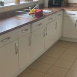 Long cream high-gloss kitchen before a respray, chrome bar handles on every door, dark walnut-effect counter, white subway tile backsplash, and a window over the black sink with pink lilies in a vase on the sill.