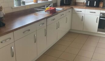 Long cream high-gloss kitchen before a respray, chrome bar handles on every door, dark walnut-effect counter, white subway tile backsplash, and a window over the black sink with pink lilies in a vase on the sill.