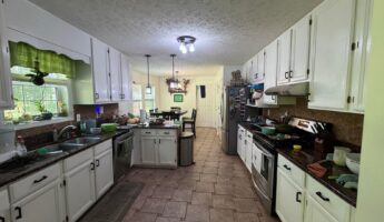 Wide 16:9 view of a long galley kitchen before a green remodel, with glossy white cabinets, a brown cork-look tile backsplash, speckled dark granite countertops, a brown basket-weave tile floor, a scalloped green valance curtain over the sink window, stainless appliances, and cluttered countertops.