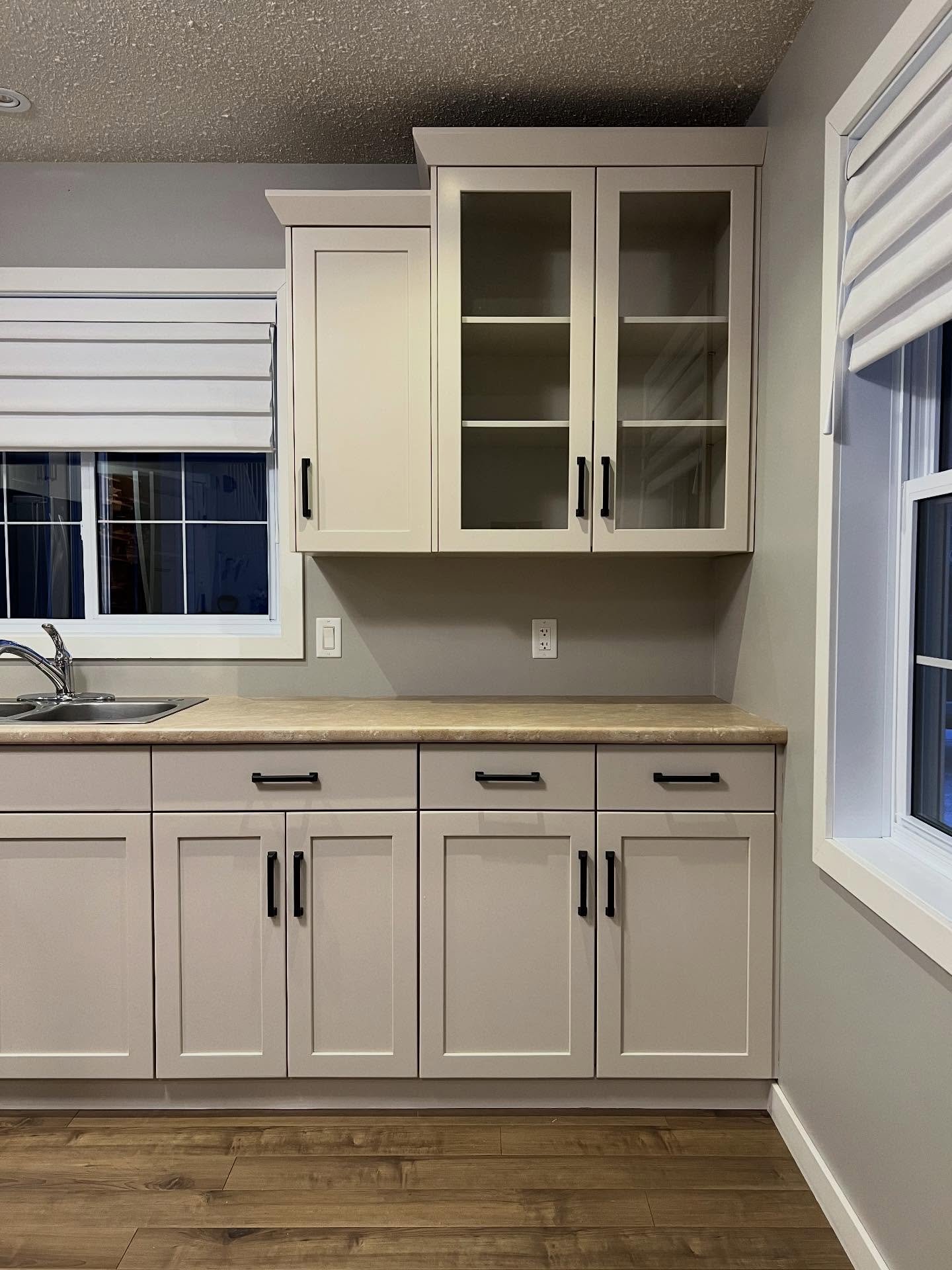 Painted glass-front display hutch in Sherwin-Williams Accessible Beige with empty glass shelves, matte black pulls, and warm wood vinyl plank flooring below.