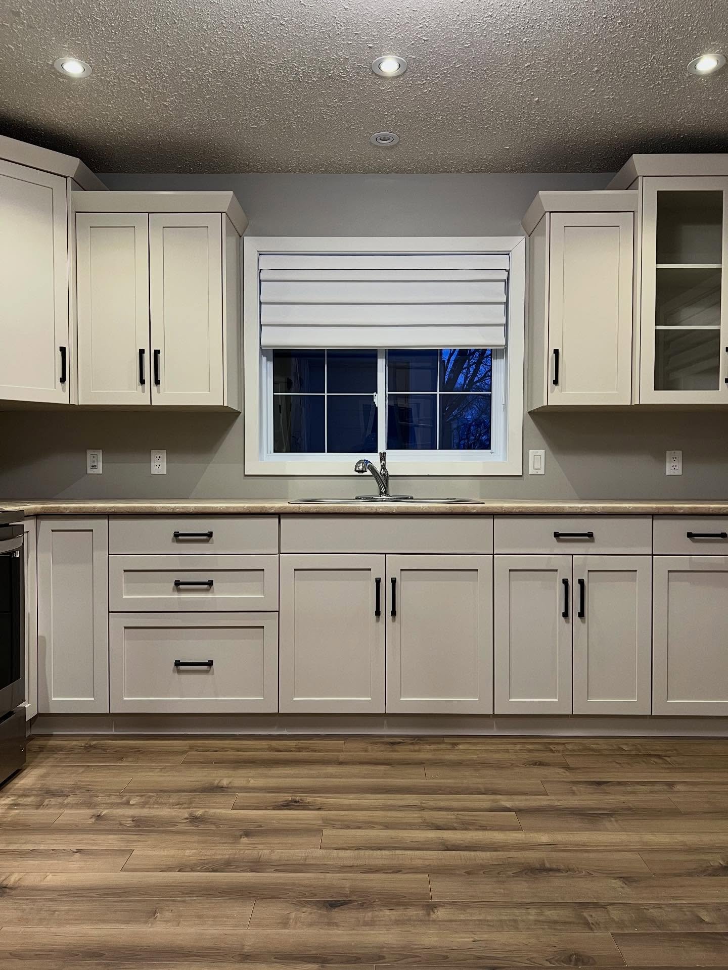 Kitchen sink wall painted Sherwin-Williams Accessible Beige at night, with a roman shade pulled up, chrome faucet, double stainless sink, tan laminate counters, matte black bar pulls, and warm wood vinyl plank flooring.