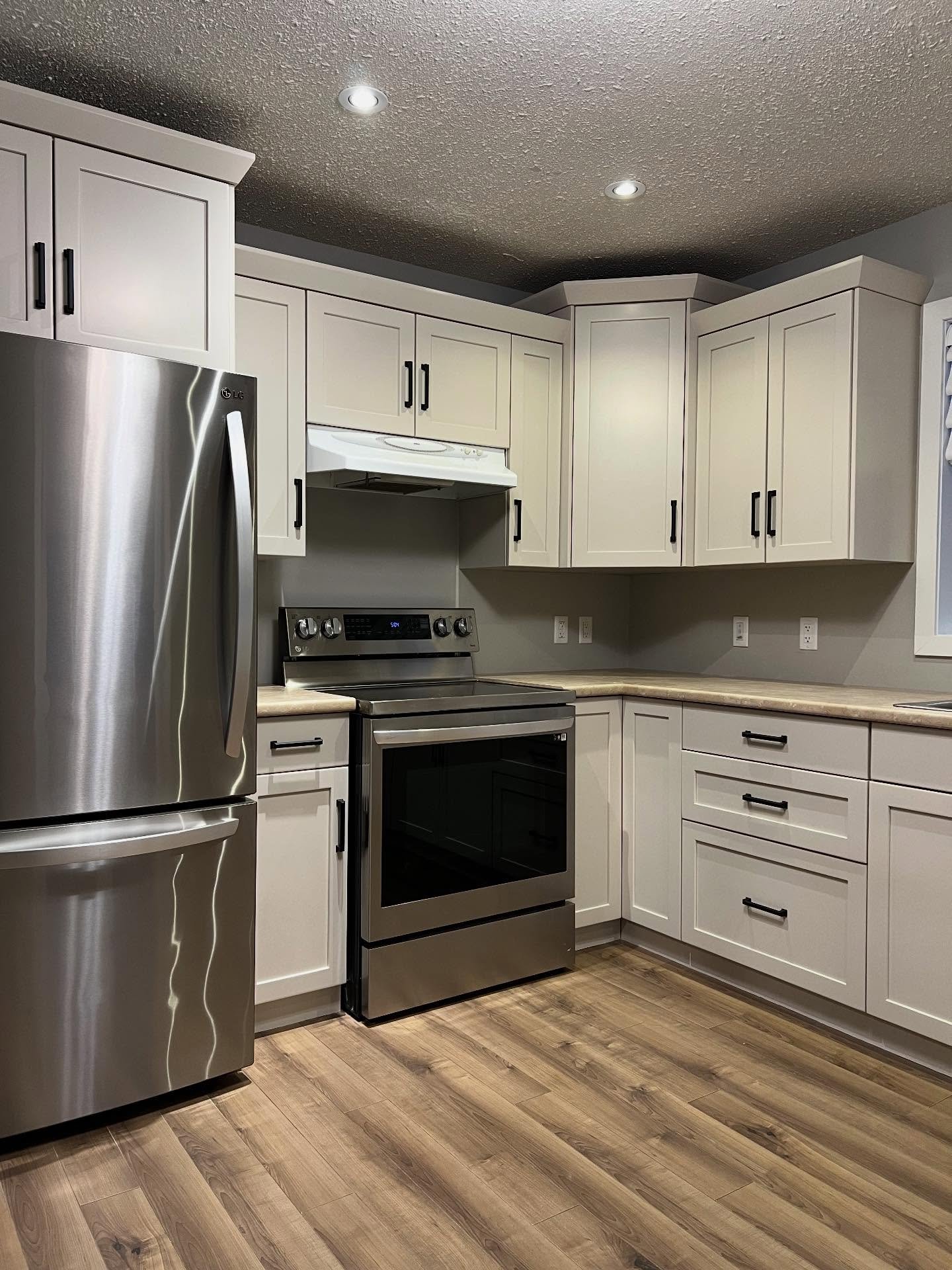 Full L-shaped kitchen painted Sherwin-Williams Accessible Beige with the same tan laminate counters, warm wood vinyl plank floor, stainless LG fridge, electric range, white range hood, and new matte black bar pulls.