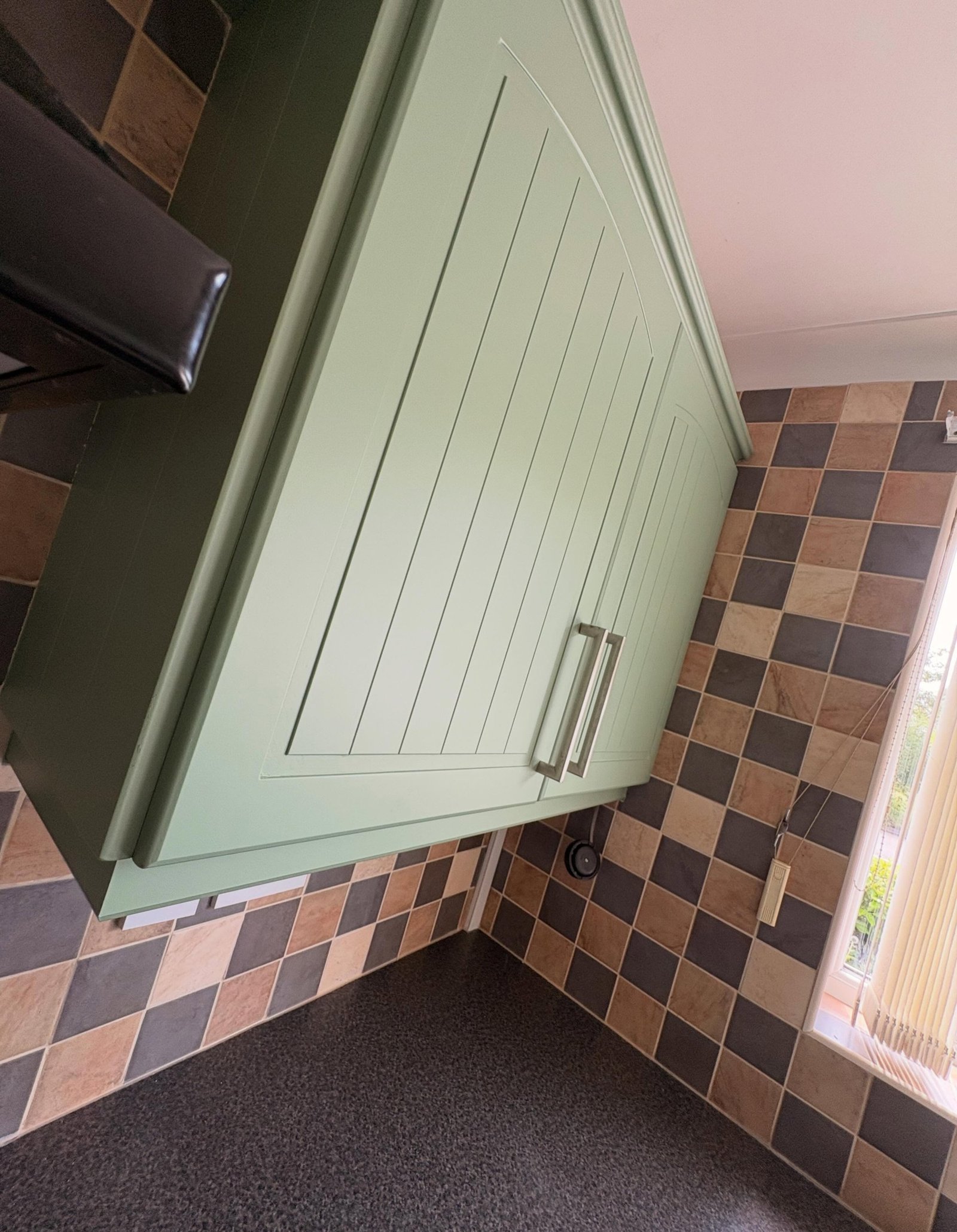 Sage green painted kitchen cabinets around a black cooker hood, with multi-tone brown and grey checkerboard tile backsplash looking intentional.