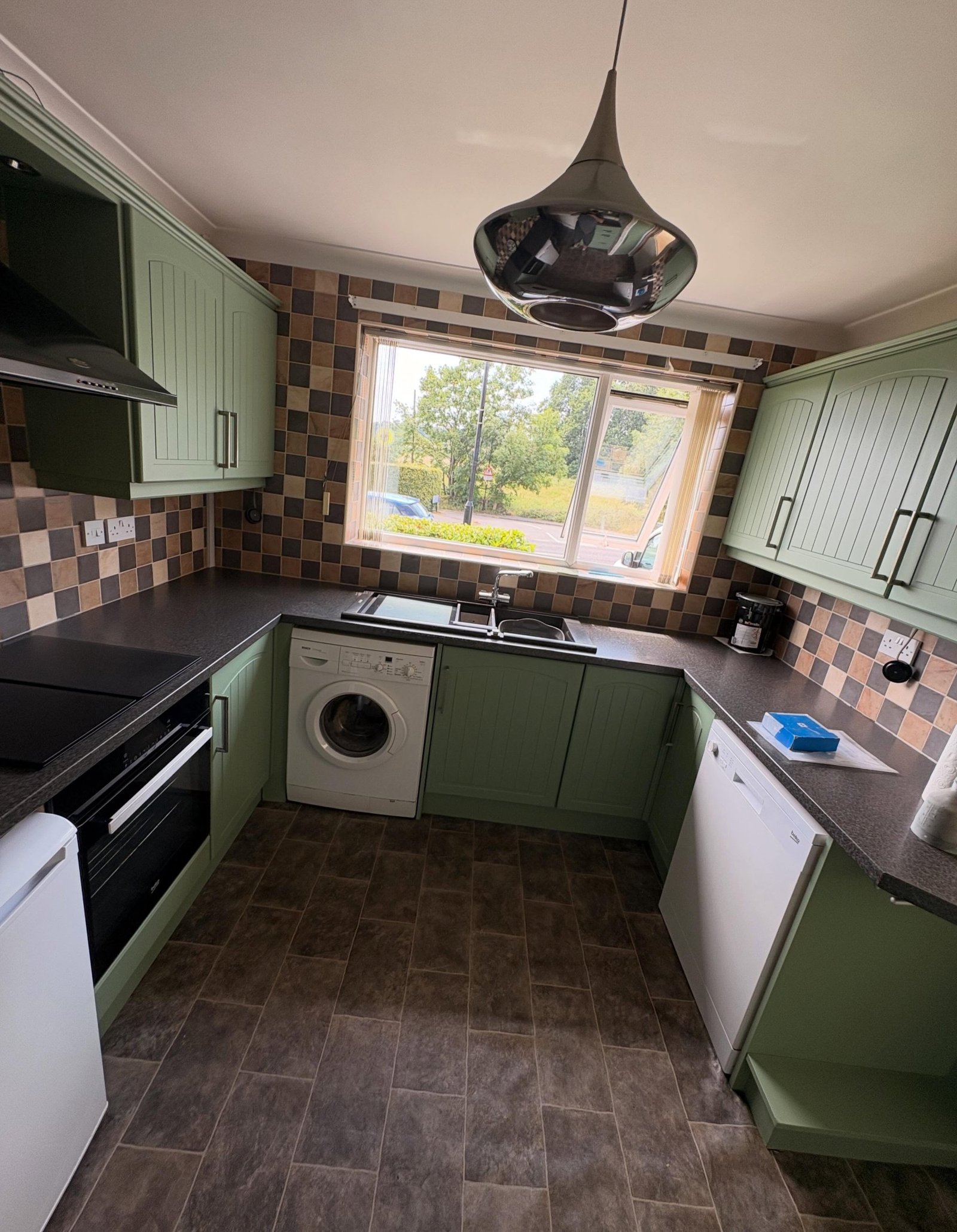 Wider view of a sage green painted kitchen oven corner with checkerboard tile, black hood and open green side shelving.