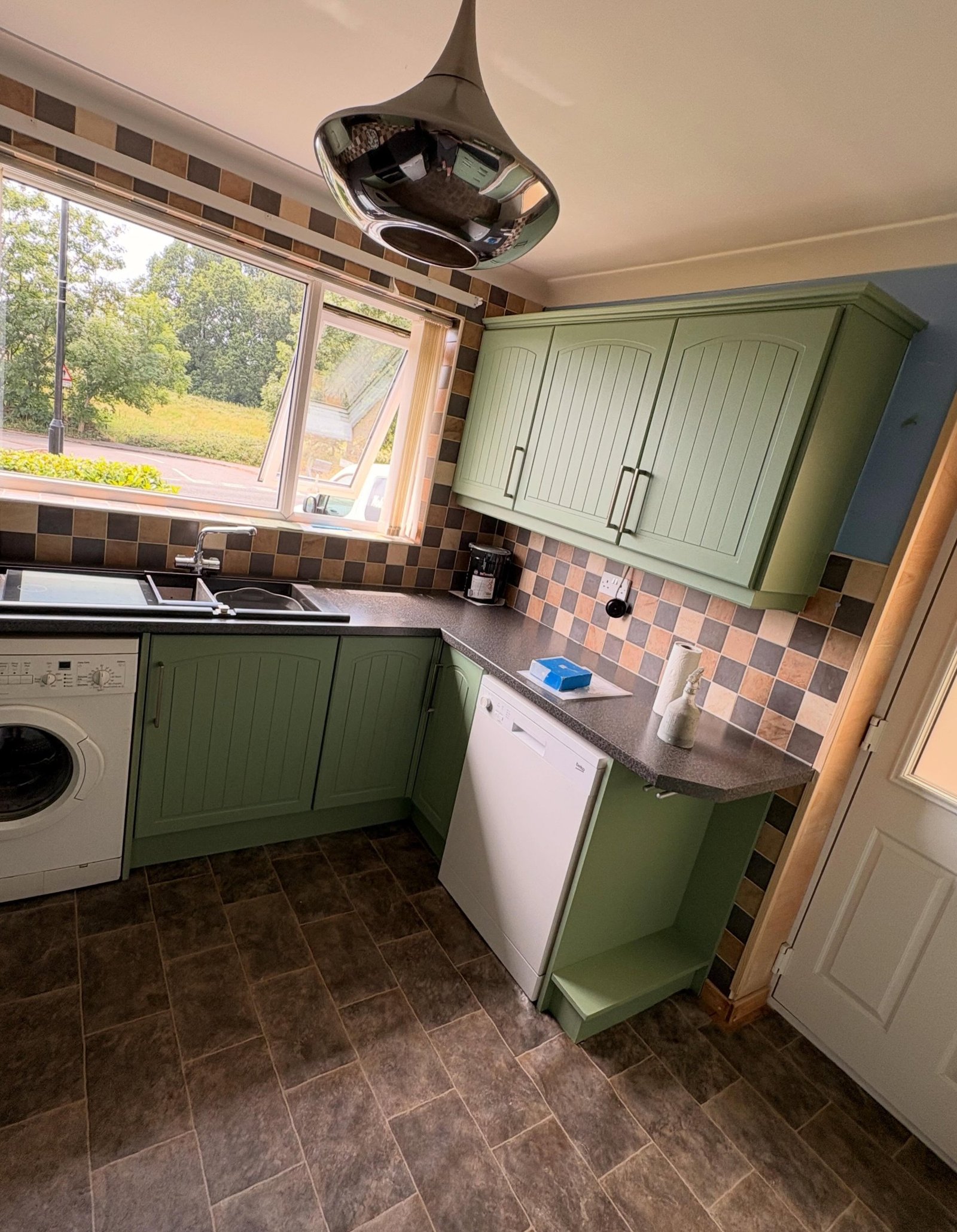 Sage green kitchen cabinets around a window and freestanding white dishwasher, patchwork tile backsplash and dark grey floor tile.