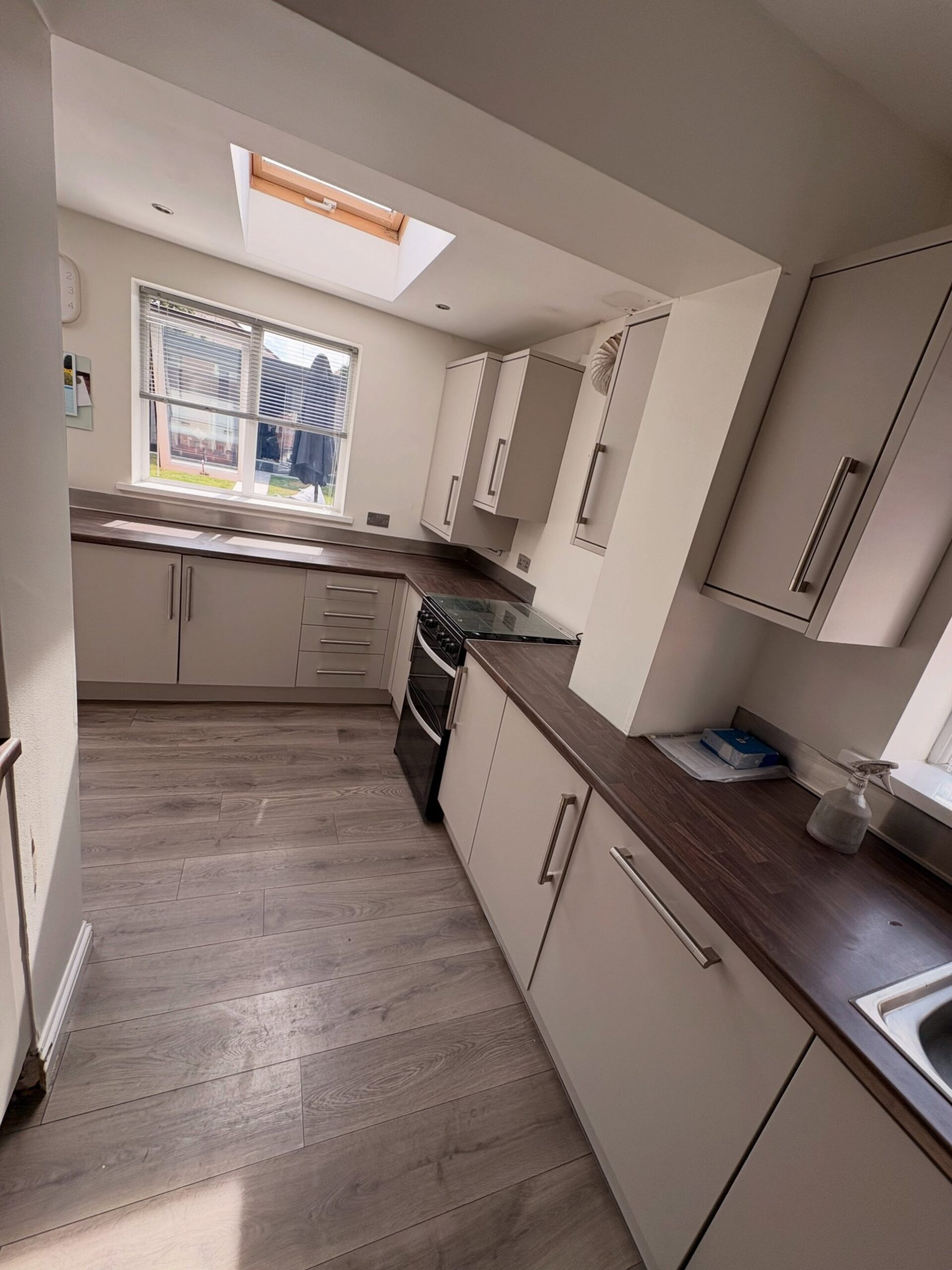 Angled view of the finished kitchen in Purbeck Stone, skylight above, dark walnut counters wrapping around to the oven and the sink, warm afternoon light on the floor.
