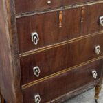 Antique four drawer wood dresser with a worn and patchy brown finish, decorative Art Deco style silver drop pulls, carved wood accents on the top drawer, and slim turned legs on small casters, standing on a concrete driveway in front of a white paneled garage door.