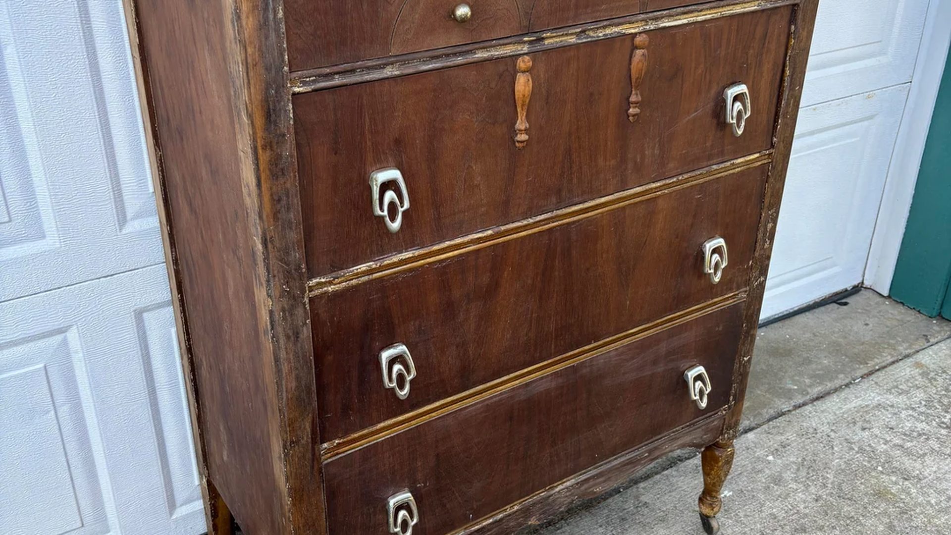 Antique four drawer wood dresser with a worn and patchy brown finish, decorative Art Deco style silver drop pulls, carved wood accents on the top drawer, and slim turned legs on small casters, standing on a concrete driveway in front of a white paneled garage door.