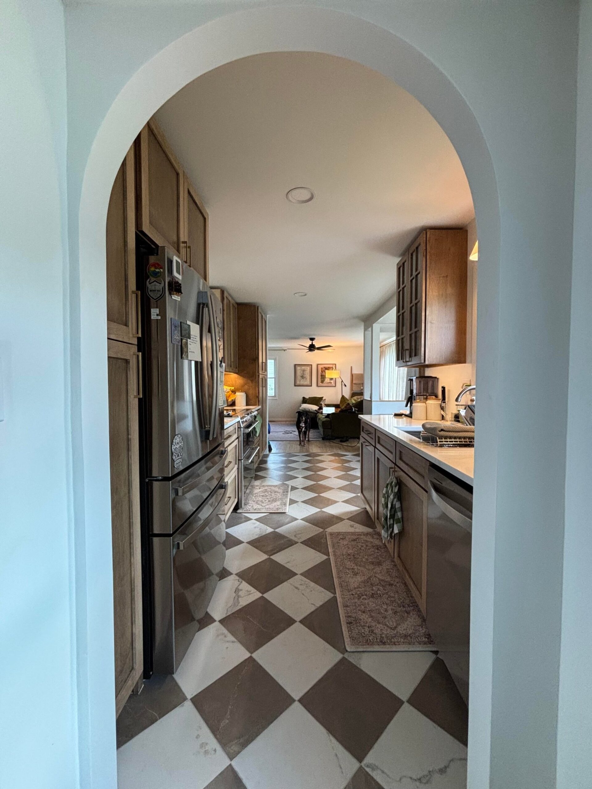 View through a round white archway into a renovated kitchen with stained wood cabinets, a stainless French-door fridge, checkered diagonal tile flooring in cream and brown, a runner rug on the floor, and a dog standing near the living area at the back.