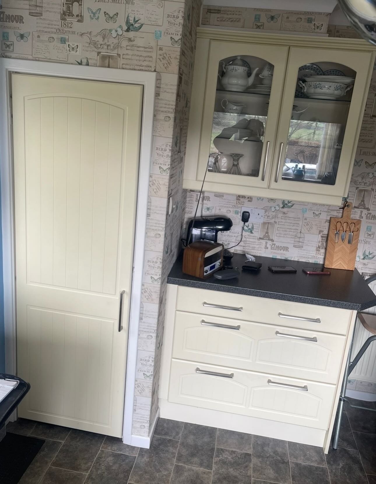 Cream tall pantry cupboard next to cream drawers, set against butterfly and vintage postcard wallpaper, with glass-front wall cabinet above.