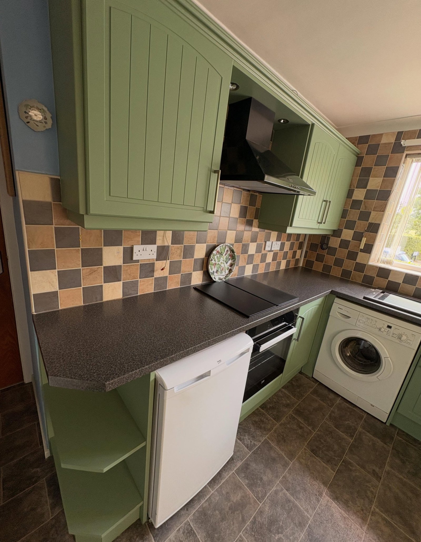 Cluttered cream kitchen corner with a small white under-counter fridge, black oven, stool, and dated patchwork tile backsplash.
