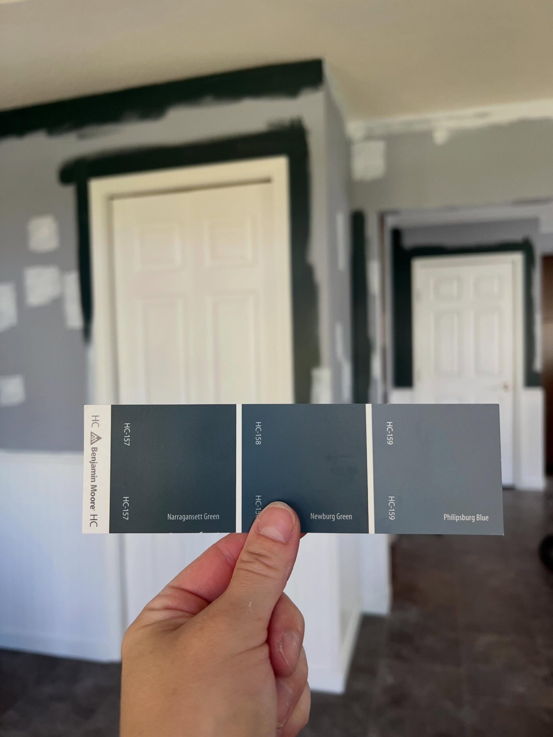 Hand holding a Benjamin Moore paint swatch card showing three Historical Collection colors, HC-157 Narragansett Green, HC-158 Newburg Green, and HC-159 Philipsburg Blue, against a wall mid-renovation with gray paint and dark green test patches around a white door and the ceiling edge.