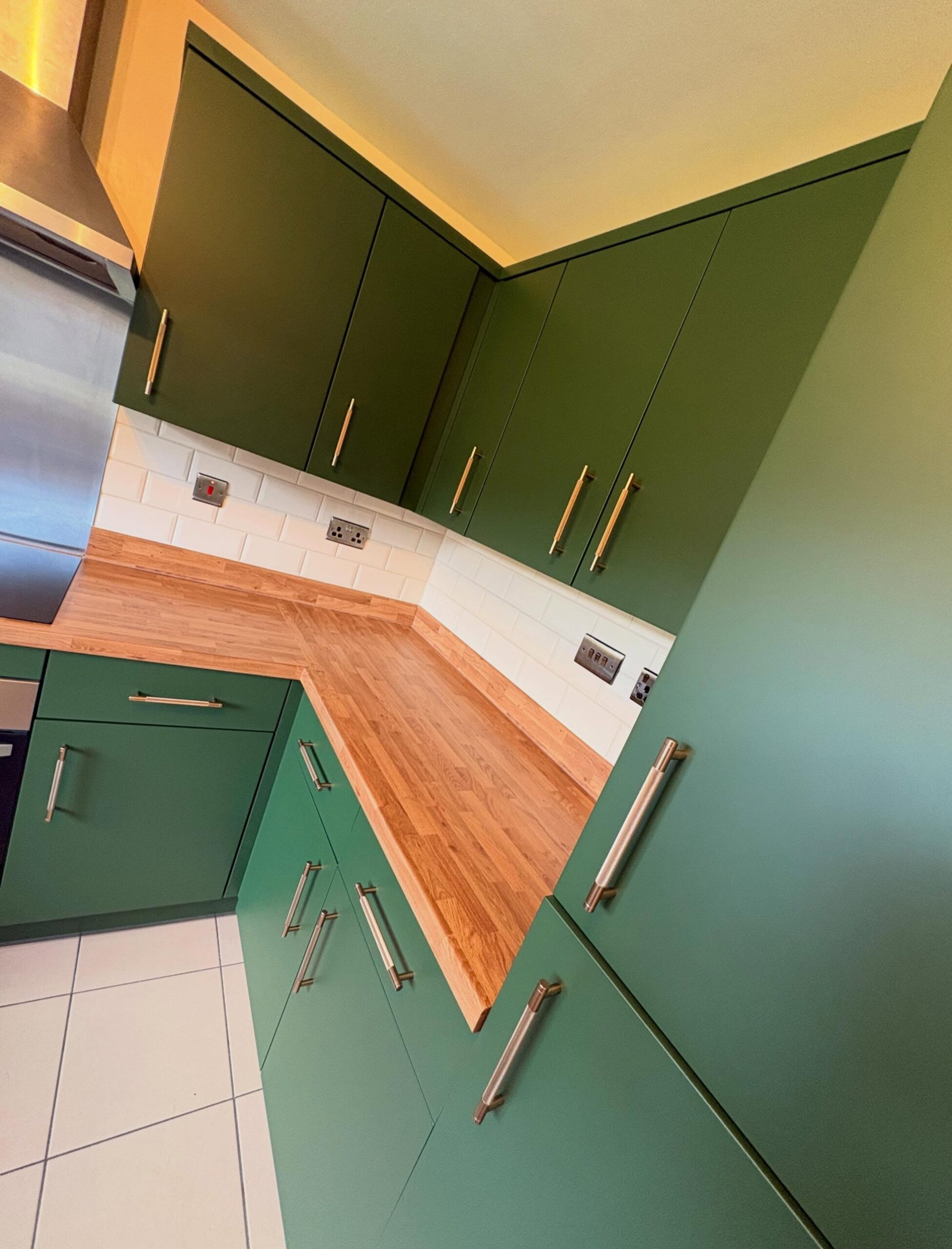 Close-up angled view of the deep green matte kitchen cabinets showing the brushed brass bar pulls, oak counter, and white subway tile detail.