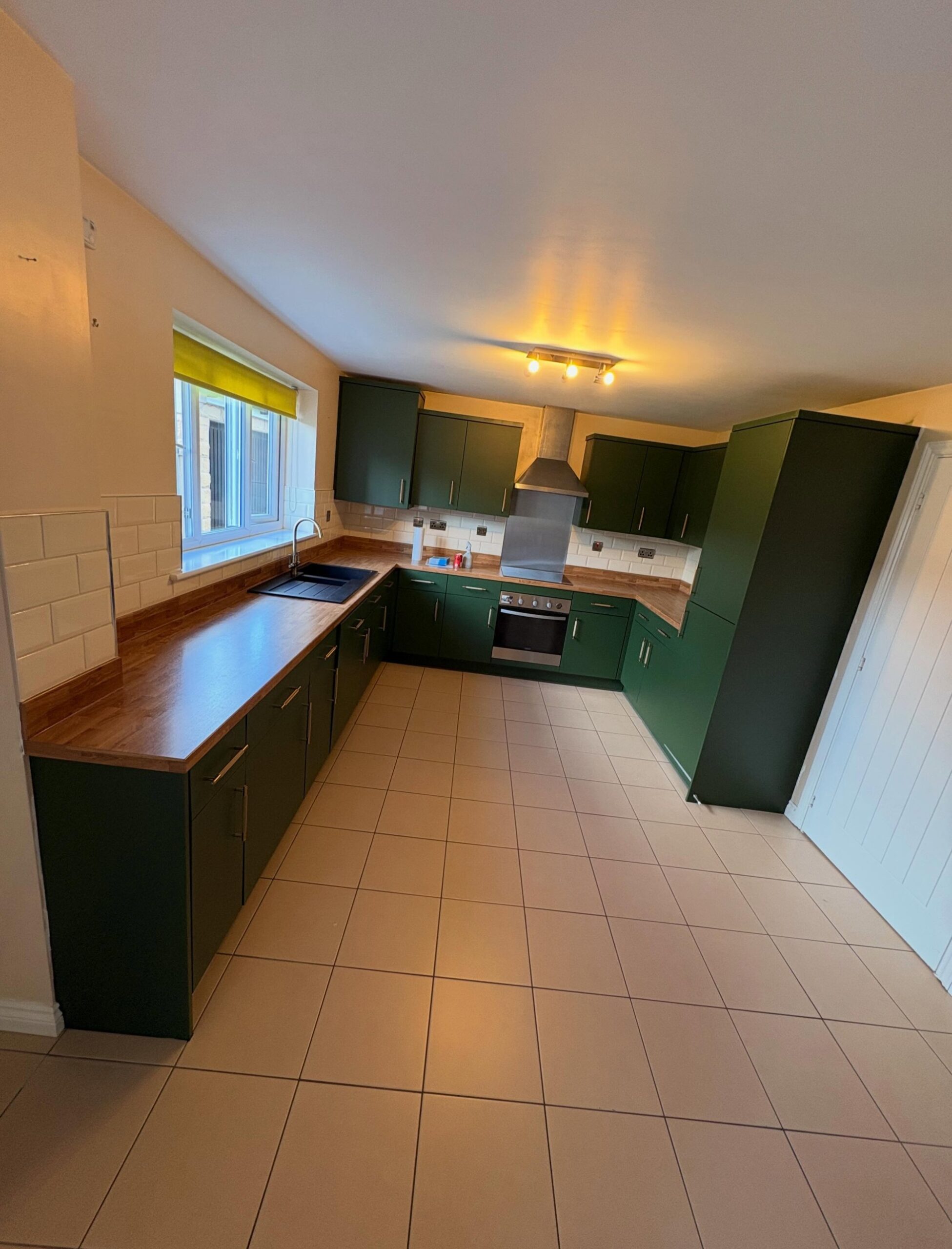 Full doorway view of the finished kitchen, deep green matte cabinets wrapping three walls in a U-shape, oak counters, cream floor tile, and a yellow shade at the window over the sink.