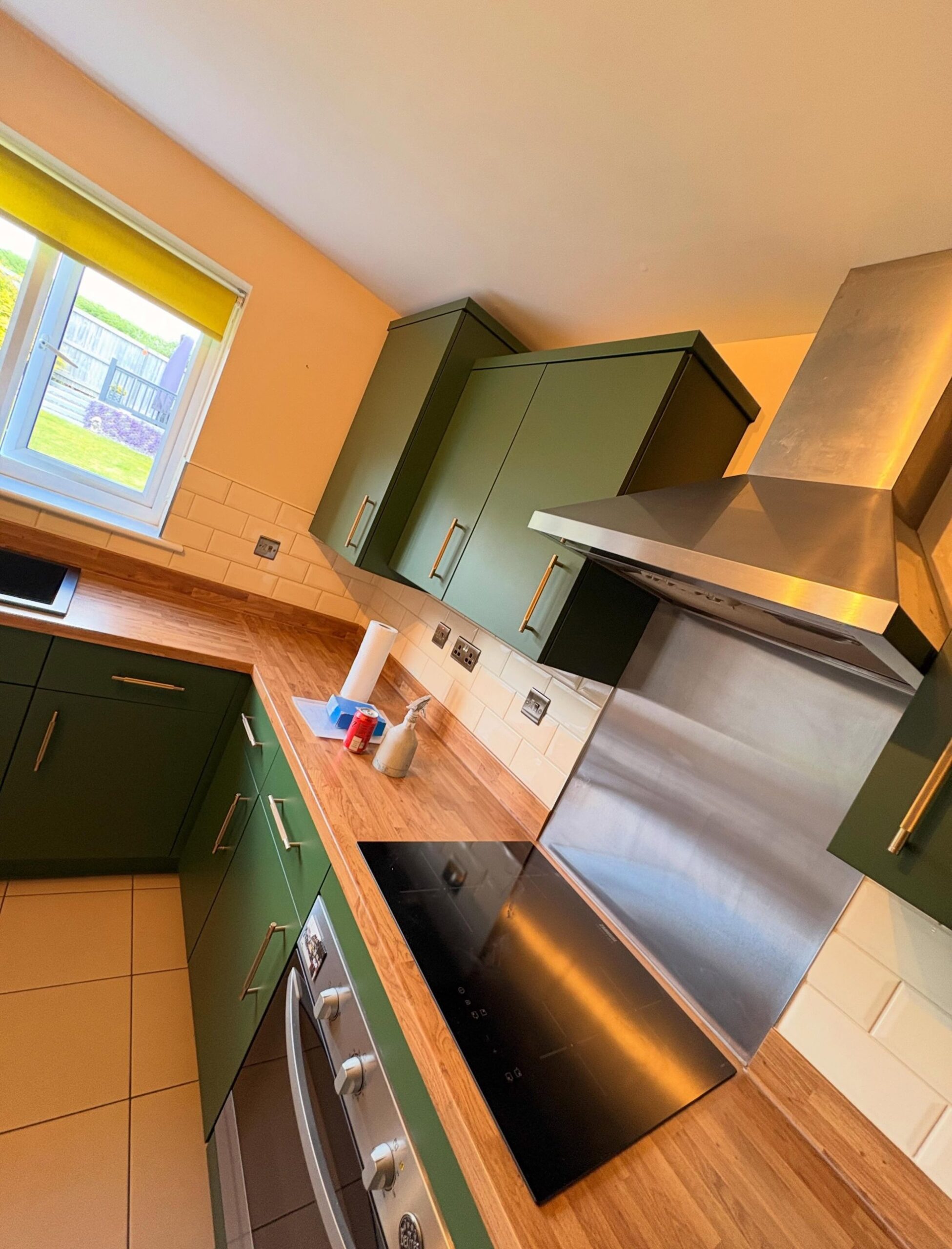 Angled view of the kitchen cooktop wall after the respray, deep green matte upper and lower cabinets with brushed brass bar pulls, oak counter, white subway tile, and a stainless steel chimney hood above the induction cooktop.