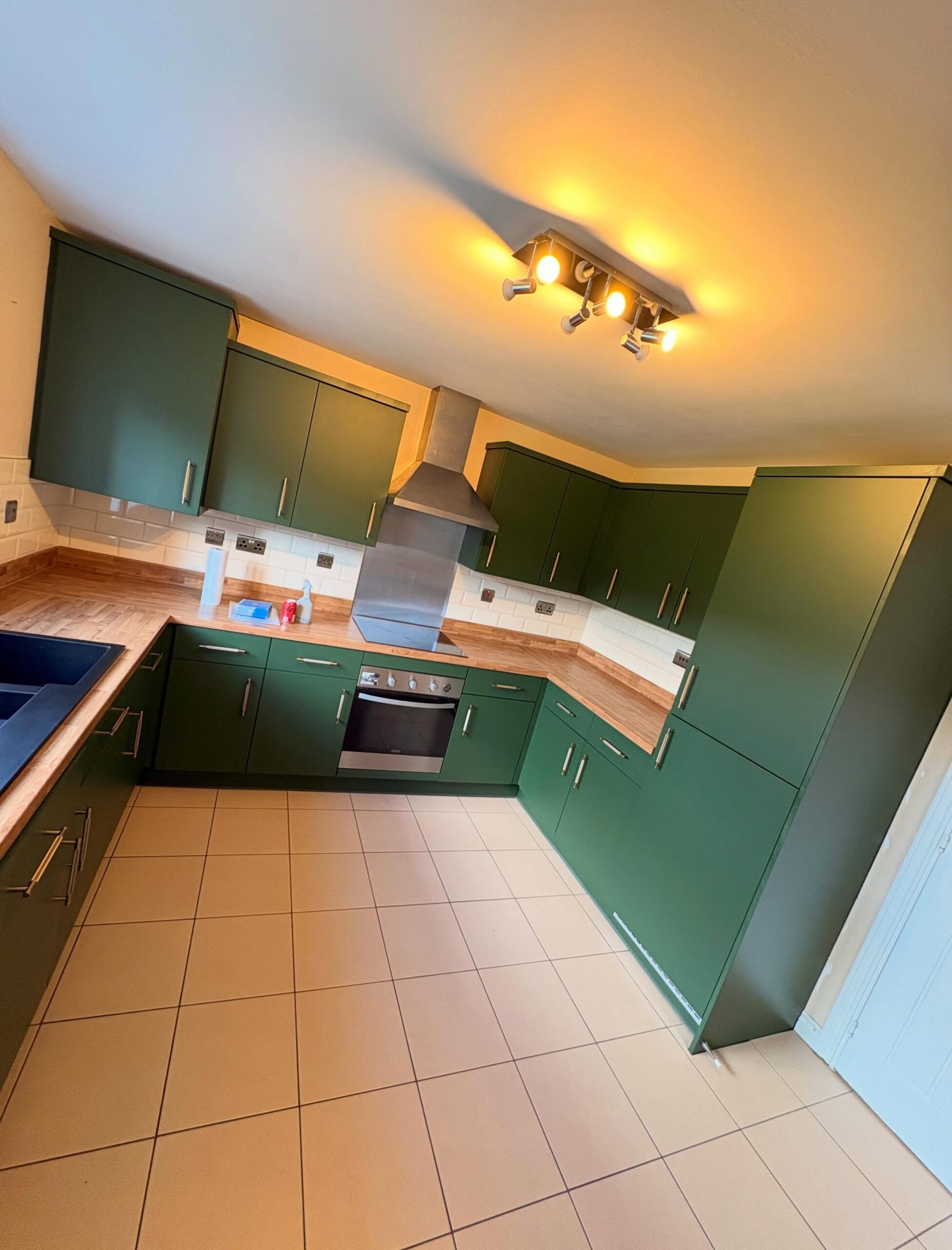 Wide angle view of the kitchen hood corner, deep green matte cabinets, oak counters, stainless chimney hood above the oven, and the ceiling spotlight bar.