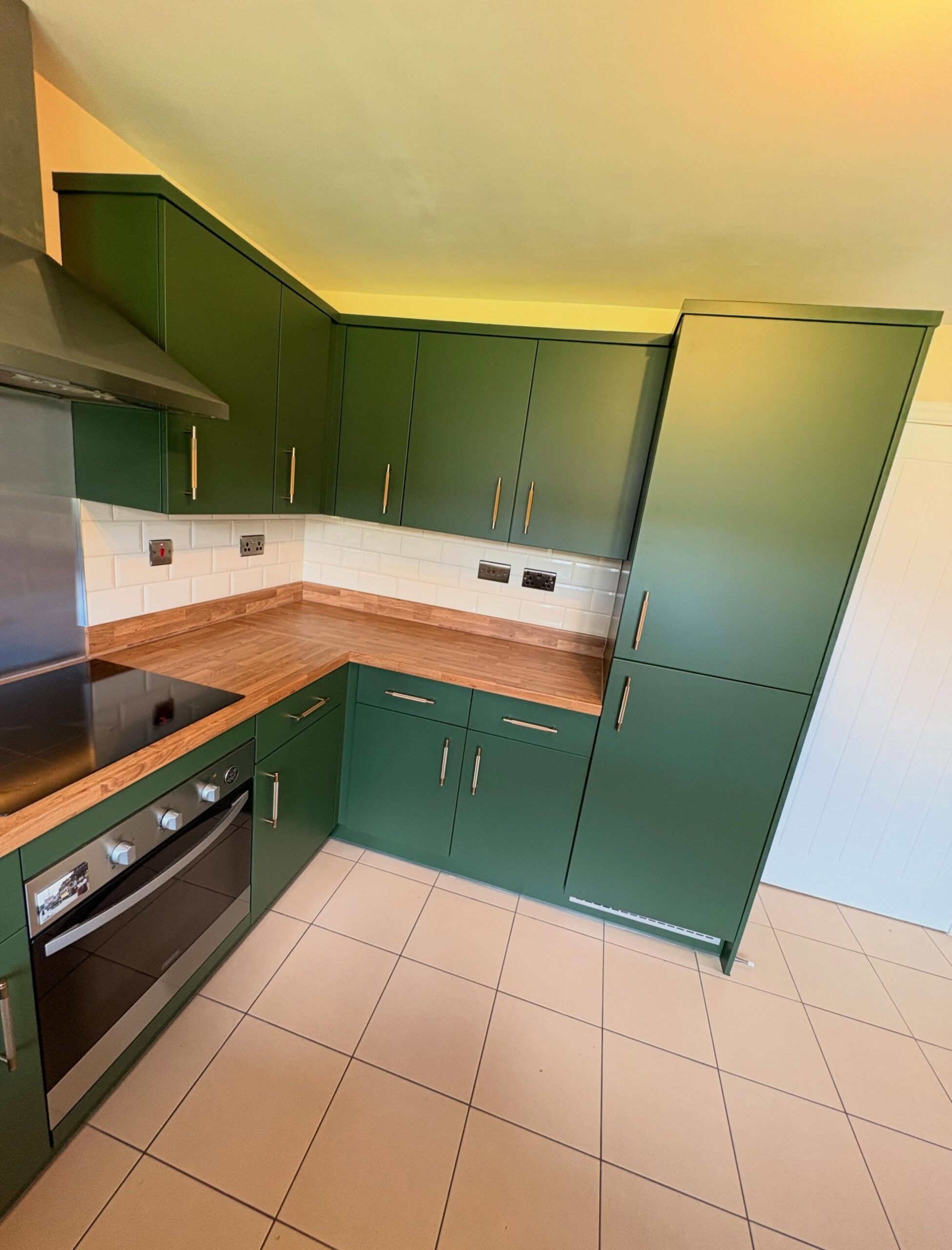Deep green matte pantry corner with full-height pantry cabinet, brushed brass bar pulls, oak counter, and white subway tile under a sloped ceiling.