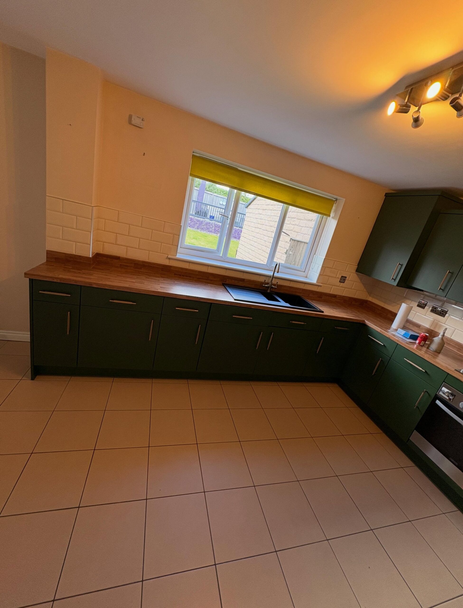 Long view of the same kitchen after the respray, cabinets now in deep Farrow and Ball Beverly Green matte, brushed brass bar pulls, light natural oak counter and the same black sink under a window with yellow shade.