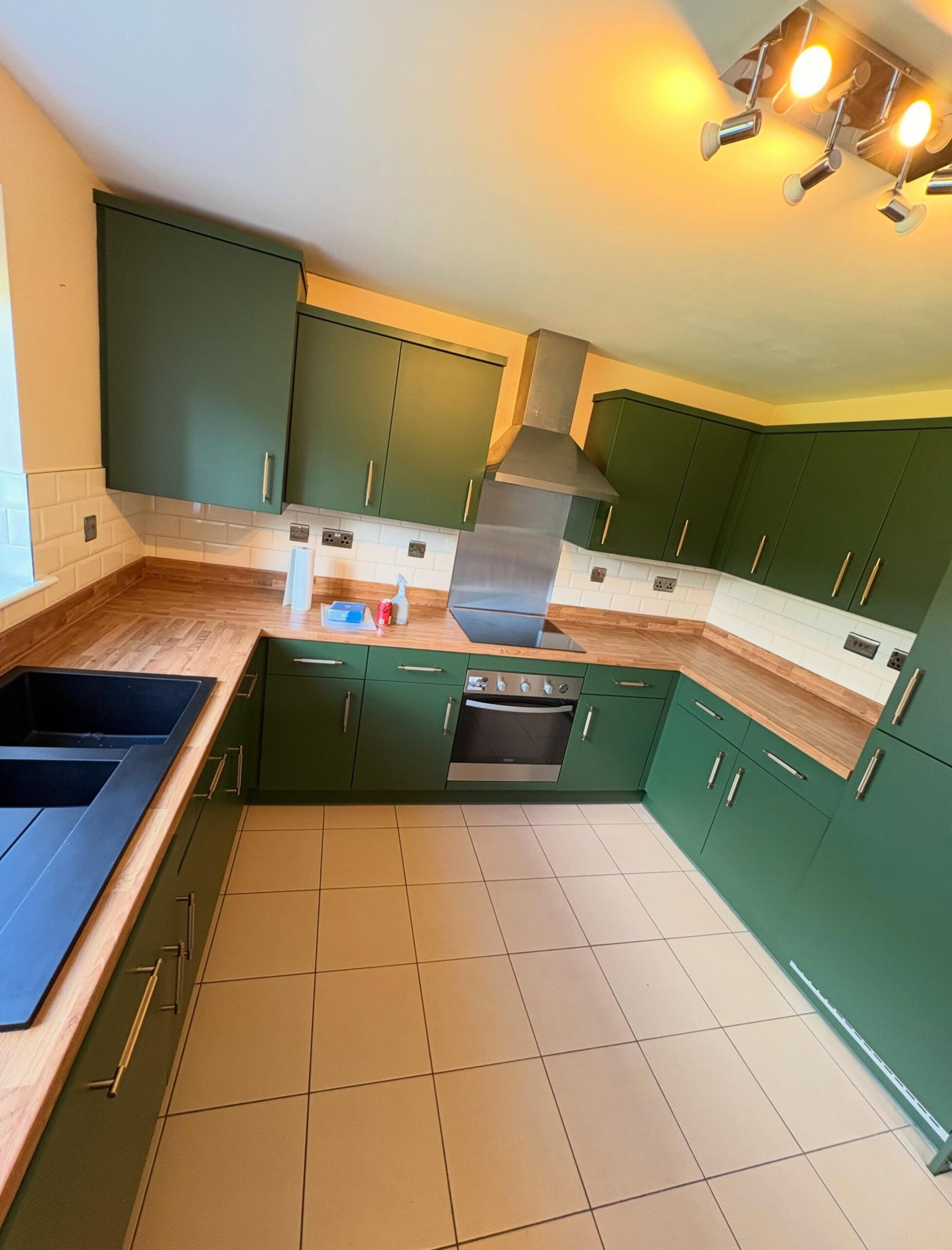 Wide view of the completed kitchen in deep Farrow and Ball Beverly Green matte, U-shape layout with oak counters, white subway tile, black granite sink on the left, oven and induction cooktop in the center, and brushed brass bar pulls throughout.