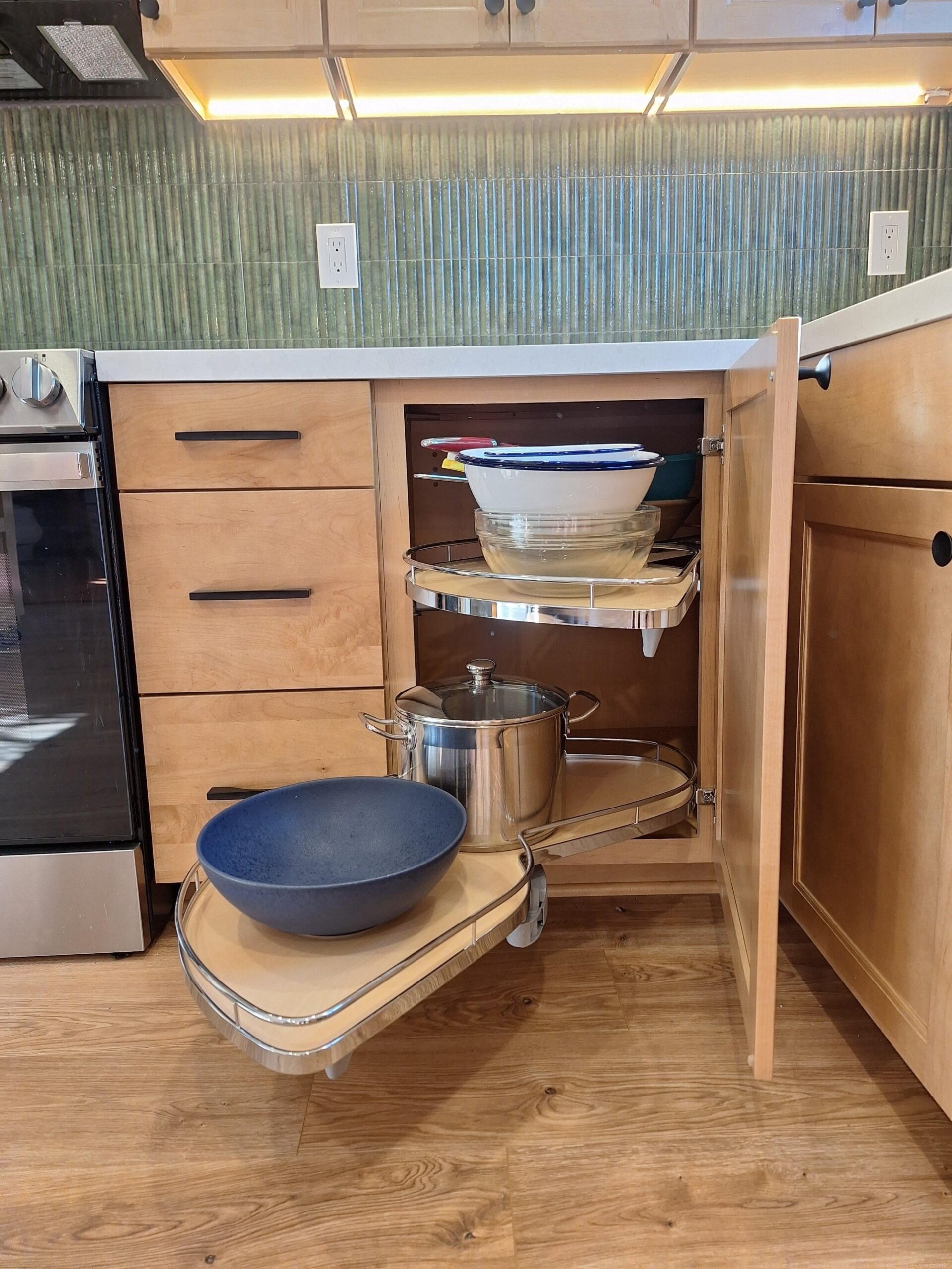 Open blind corner lower cabinet with a two-tier peanut-shaped pull-out shelf swung out fully, holding mixing bowls, a stainless stockpot, and a dark blue serving bowl.