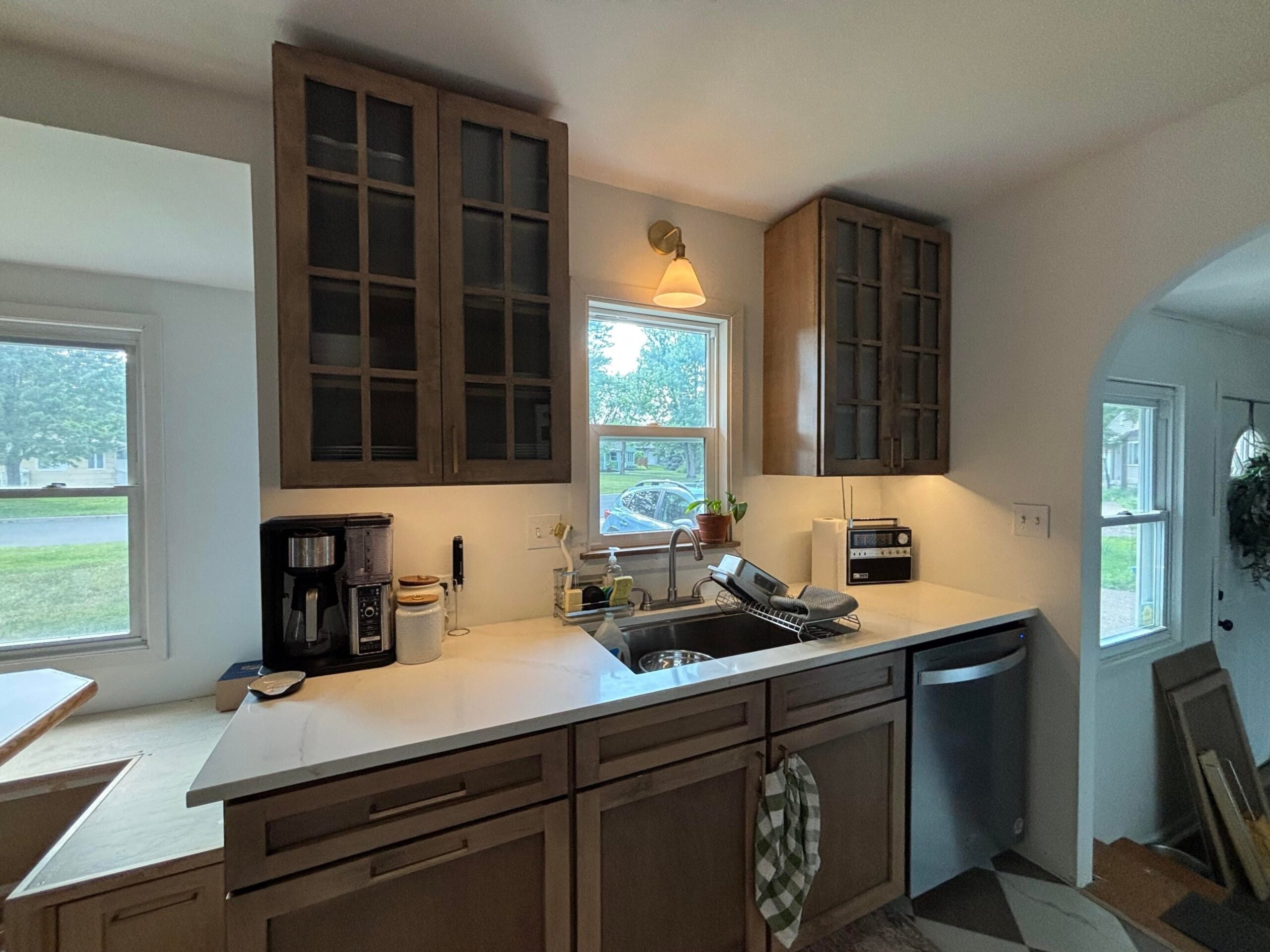 Kitchen sink area with stained wood upper cabinets with multi-pane glass fronts, a brass wall sconce with a white cone shade, a drip coffee maker and a canister on the quartz counter, a potted plant on the window ledge, and a dishwasher at the right.