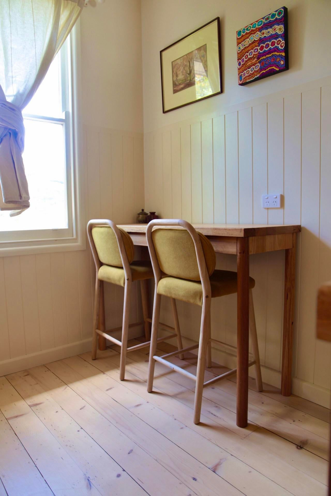 Small wood breakfast bar against a white paneled wall with two mustard upholstered stools, framed art above, and a sunny window beside it.