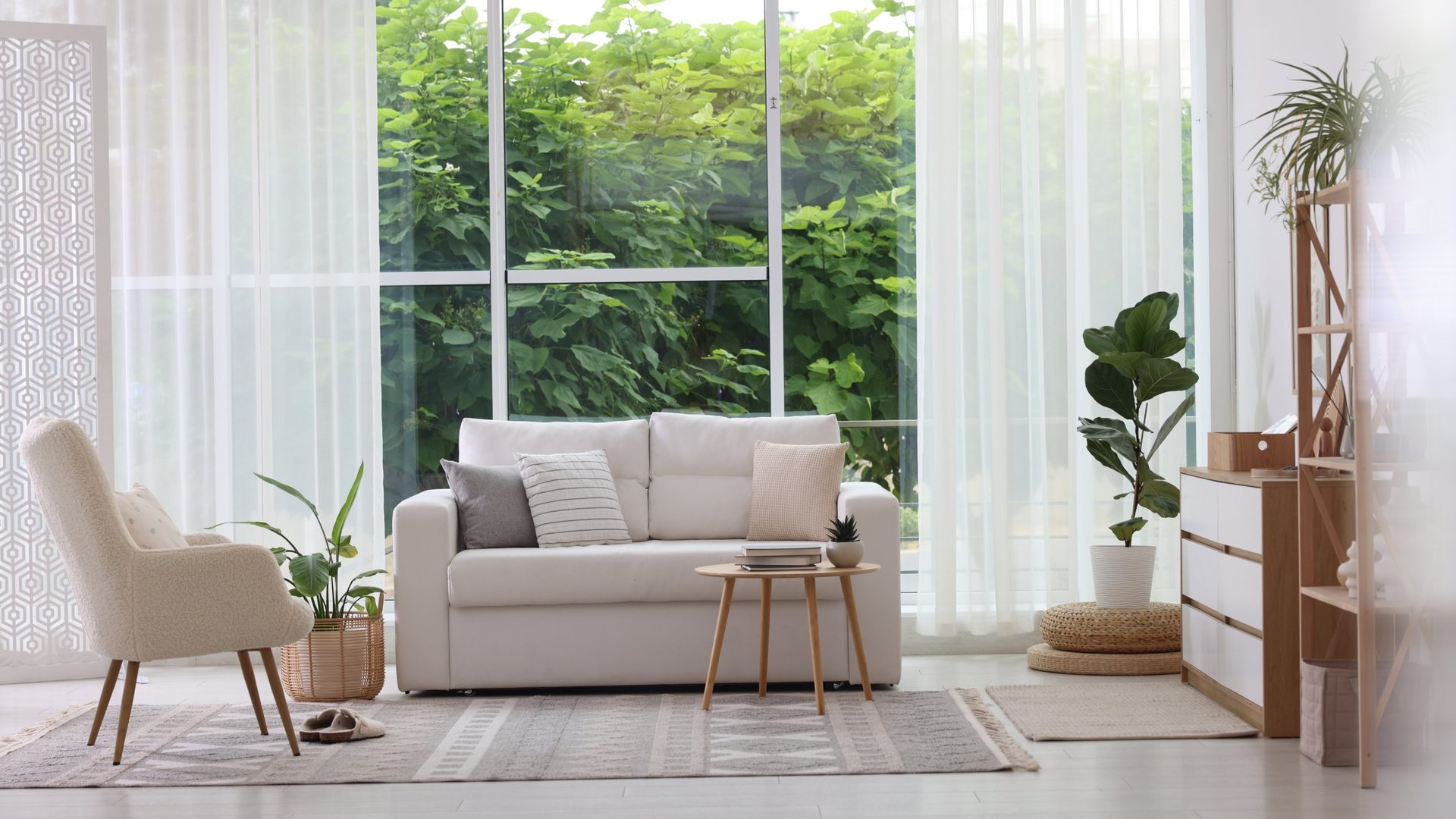 Bright Scandinavian living room with a white two-seat sofa dressed in neutral mix-and-match cushions, a cream boucle accent chair on tapered wood legs, a small round wood side table, a geometric print flat-weave rug, a potted fiddle leaf fig and traveler palm beside floor-to-ceiling windows draped in sheer white curtains overlooking lush green trees, and a light wood ladder shelf with white drawers on the right.