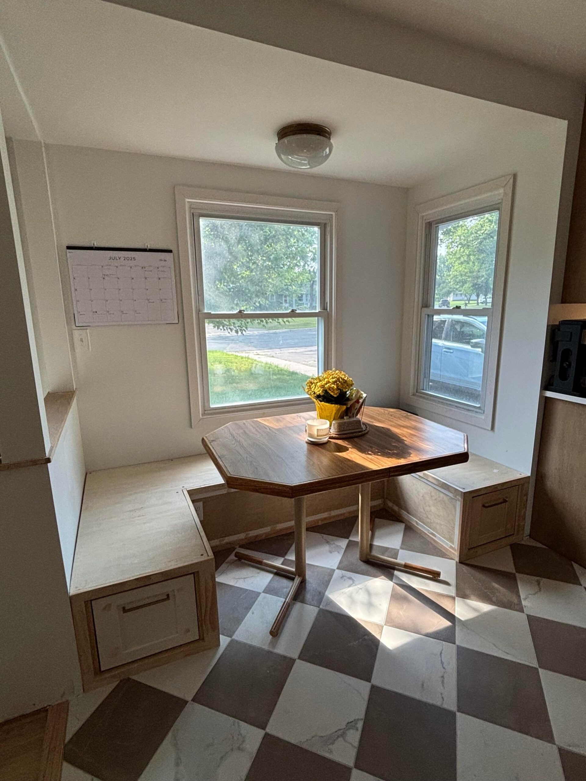 Breakfast nook with a built-in L-shaped wood storage bench with drawer fronts at the base, the same octagon wood dining table from the before shot, a yellow floral centerpiece, two tall windows looking at the street, and a July 2025 calendar on the wall.